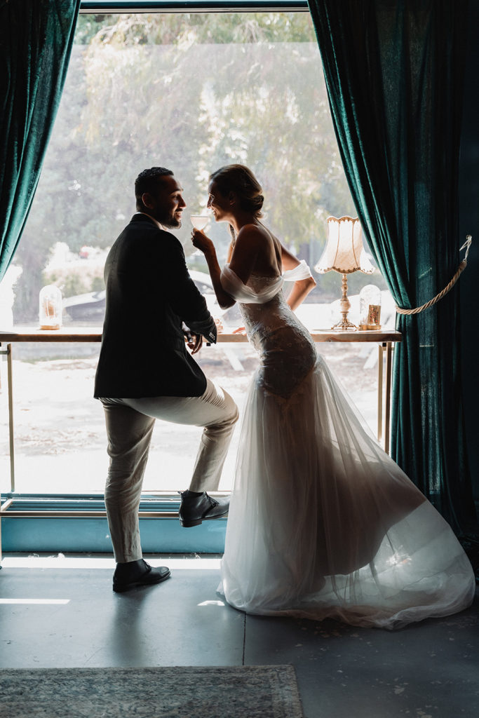newlywed couple enjoy flirtatious cocktails in front of a window wearing a stunning wedding gown and tailored suite